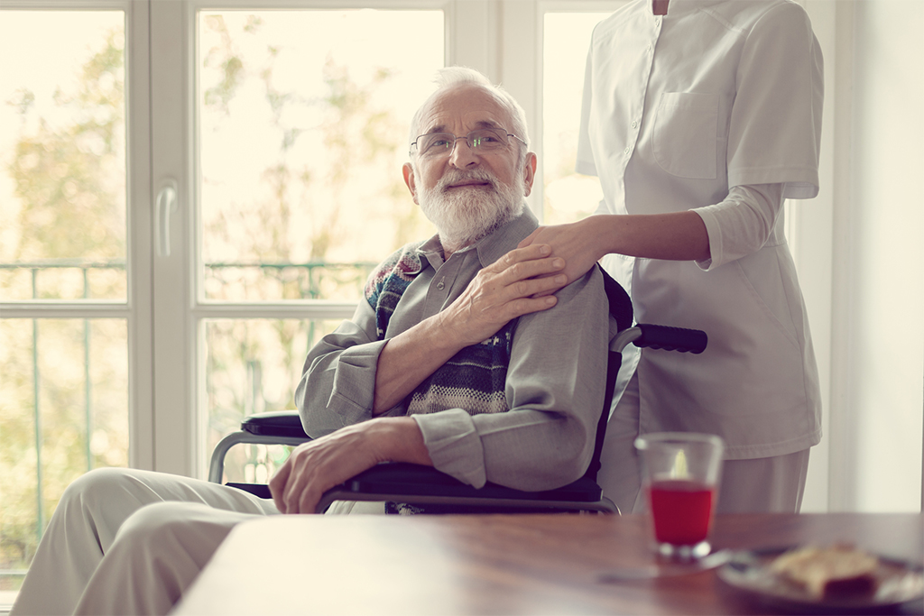 An older man in a wheelchair beside a companion caregiver at a sunlit table